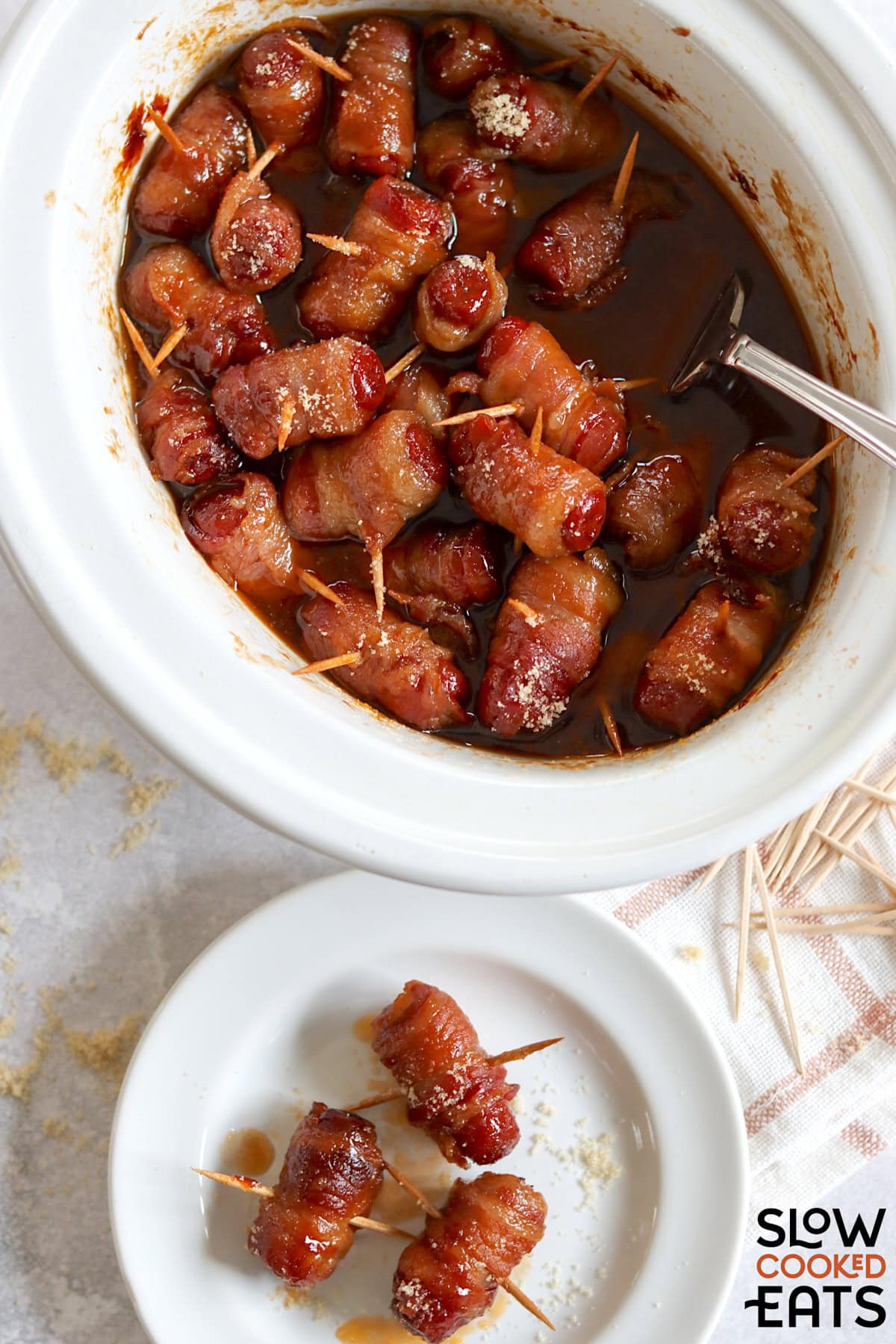 Crockpot little smokies wrapped in bacon in a white oval crockpot and three individual on a white plate.