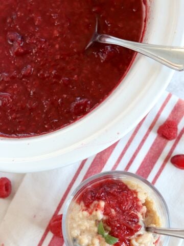 Raspberry cranberry sauce crockpot recipe in a white slow cooker, served on top of rice pudding in a glass and silver spoon.