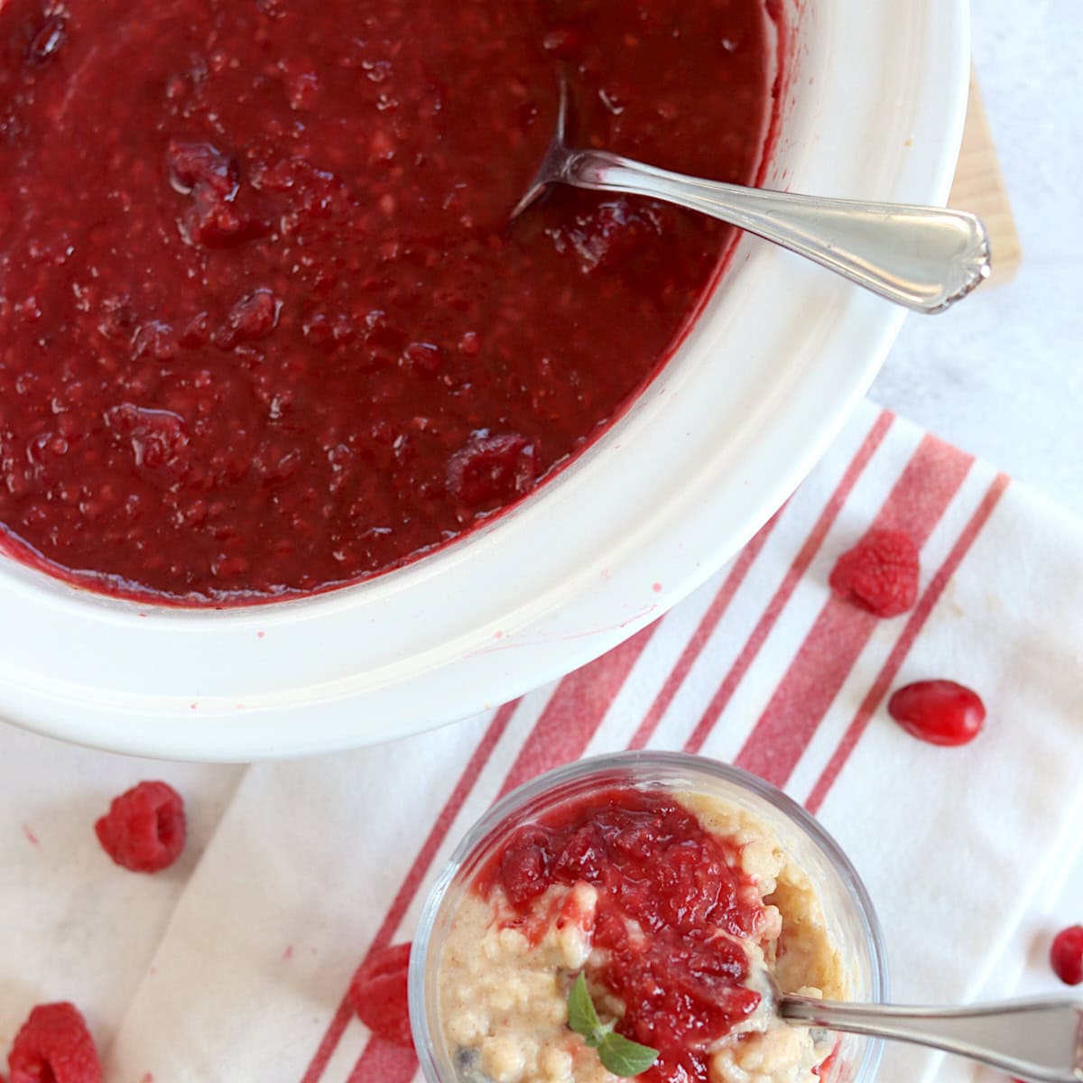 Raspberry cranberry sauce crockpot recipe in a white slow cooker, served on top of rice pudding in a glass and silver spoon.