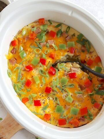 Ham and egg crockpot breakfast casserole in a white oval slow cooker and black spoon.