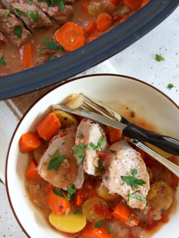 Ranch crockpot pork tenderloin with vegetables in a white bowl with silver fork and knife.
