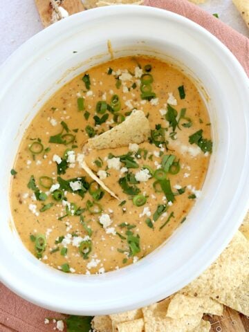 Slow cooker queso Blanco dip in a white oval crockpot and garnished with green onion and extra cheese.