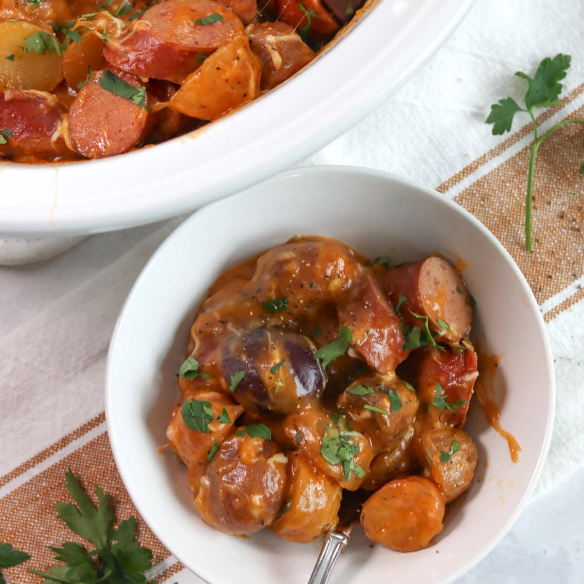 Crockpot sausage and potatoes in a white round bowl and silver spoon.