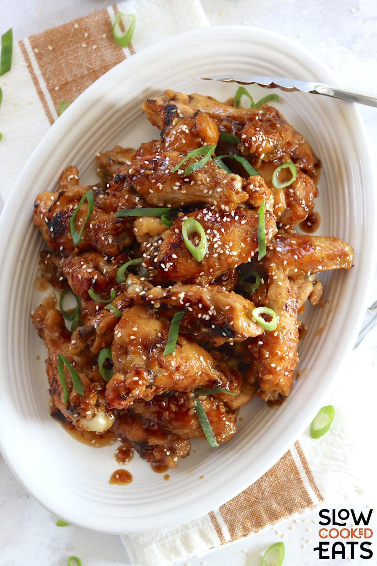 Crockpot teriyaki chicken wings on a white oval platter and garnished with green onion and sesame seeds.