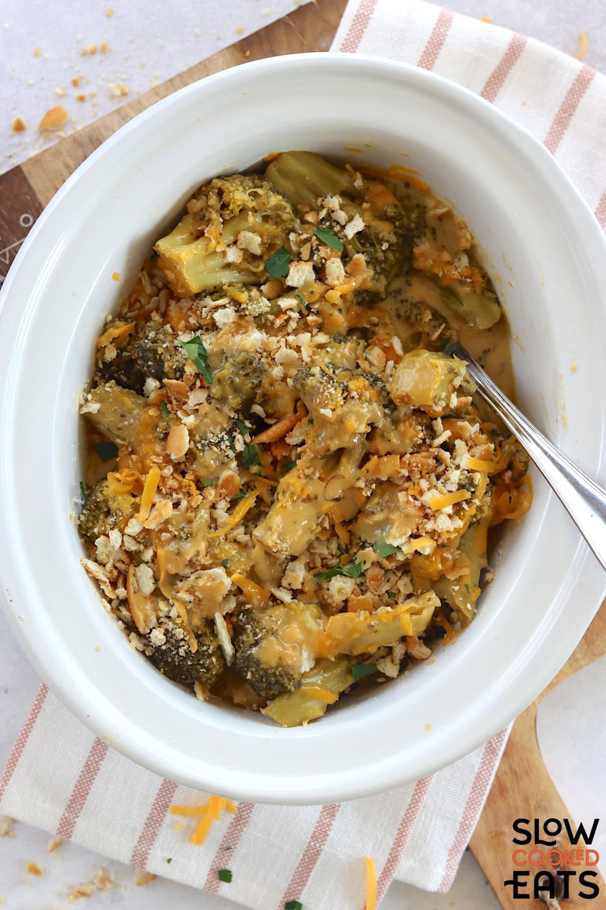 A white oval slow cooker filled with crockpot broccoli cheese casserole and a silver serving spoon.
