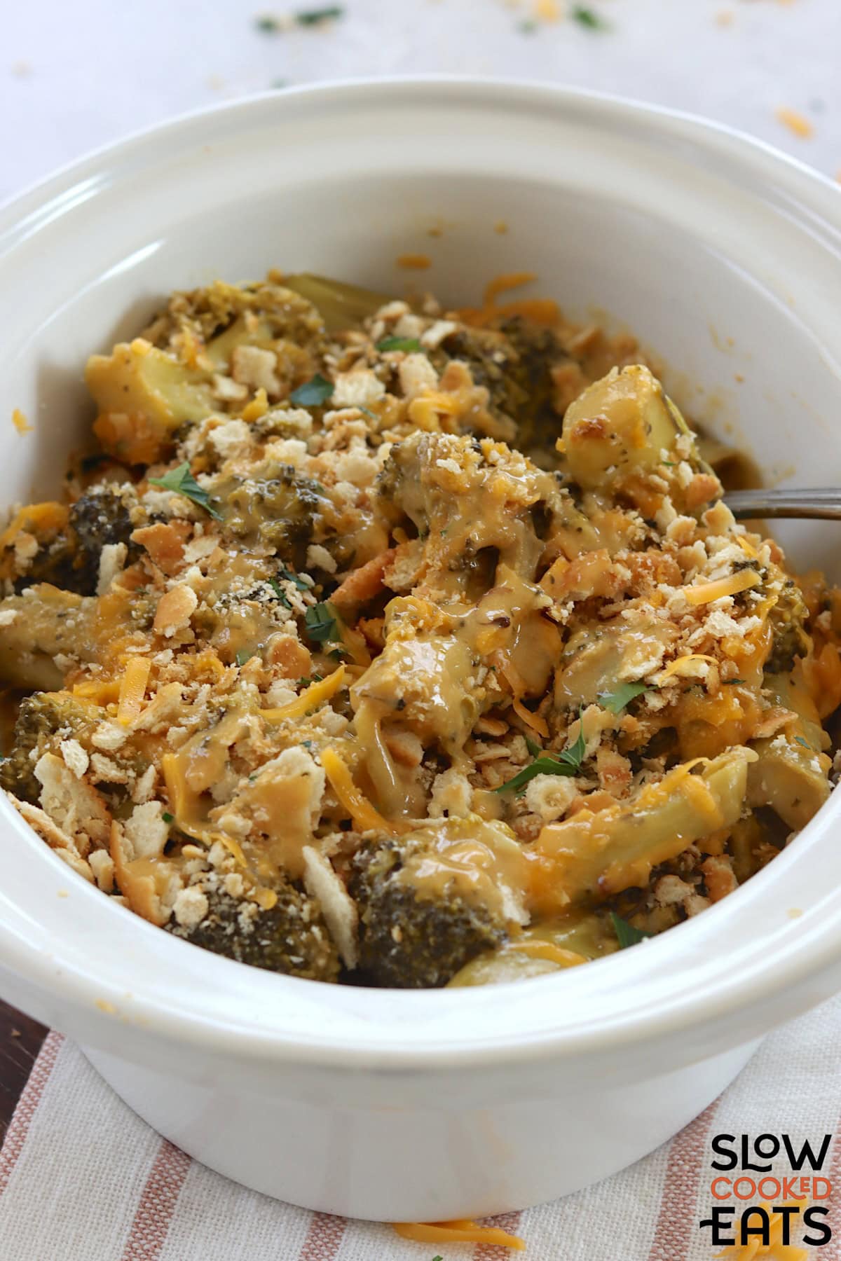 A white oval slow cooker filled with crockpot broccoli cheese casserole and a silver serving spoon.