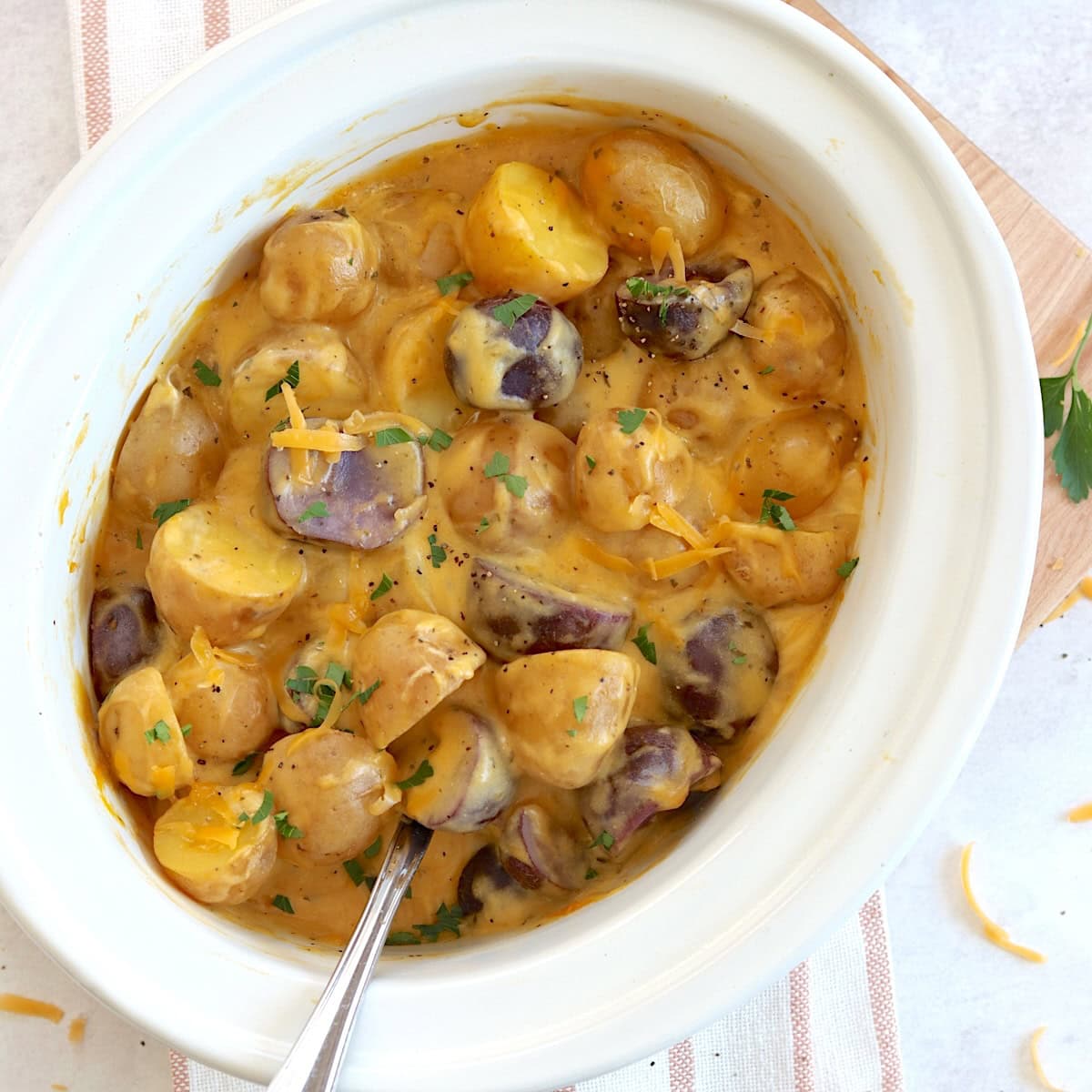 Crockpot cheesy potatoes in a white oval slow cooker and large silver serving spoon.