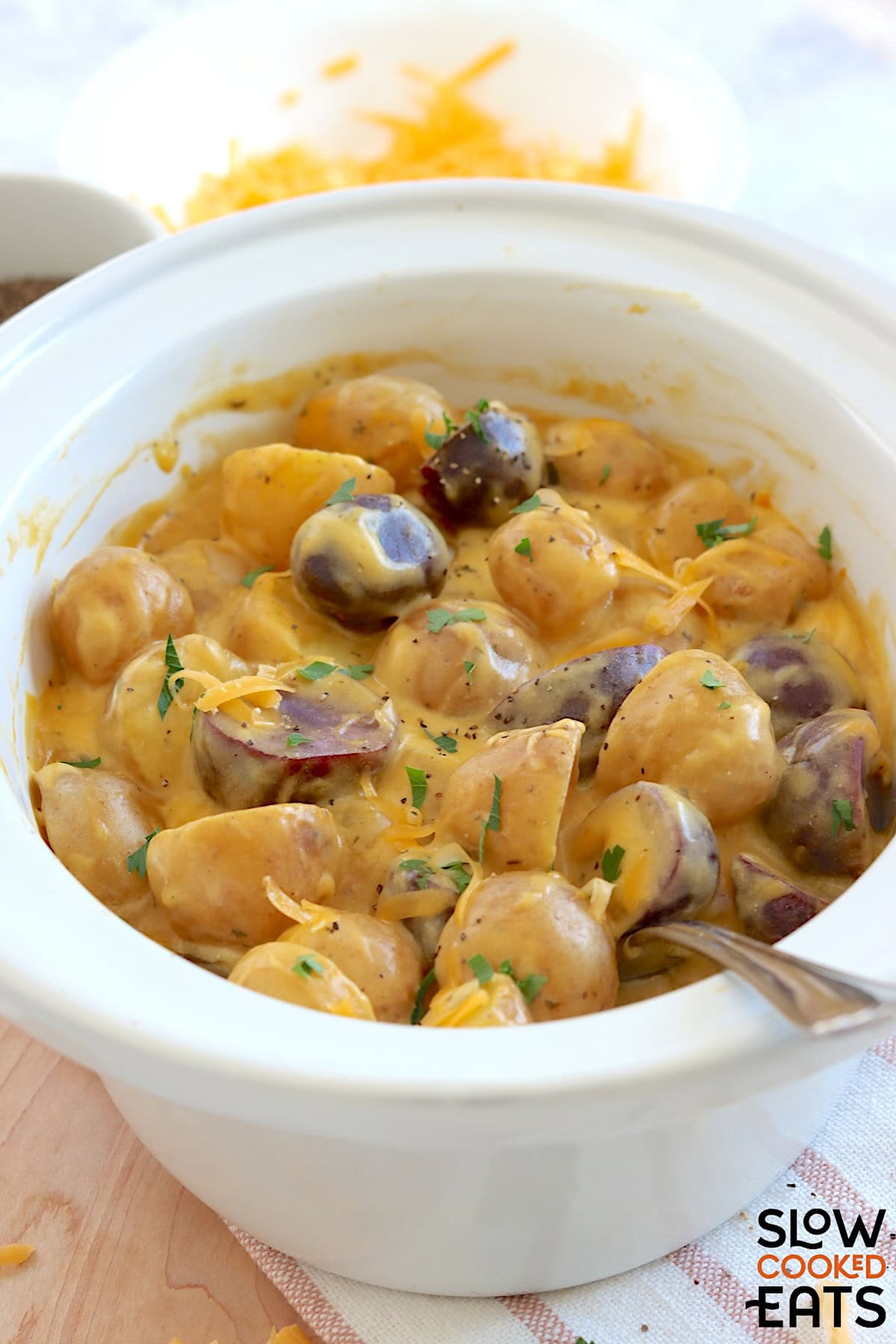 Crockpot cheesy potatoes in a white oval slow cooker and large silver serving spoon.