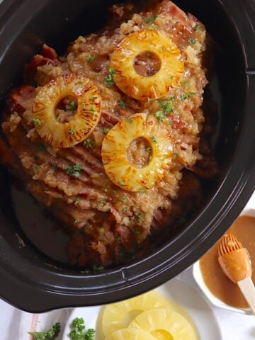 A black oval slow cooker filled with pineapple crockpot ham and topped with three seared pineapple slices.