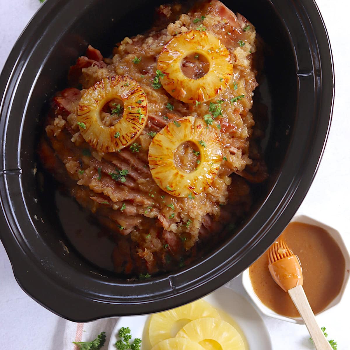 A black oval slow cooker filled with pineapple crockpot ham and topped with three seared pineapple slices.