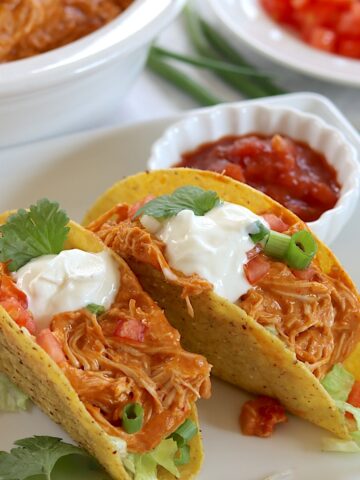 Two hard shell tacos filled with shredded queso chicken, topped with sour cream, tomatoes, and green onions on a white square plate.