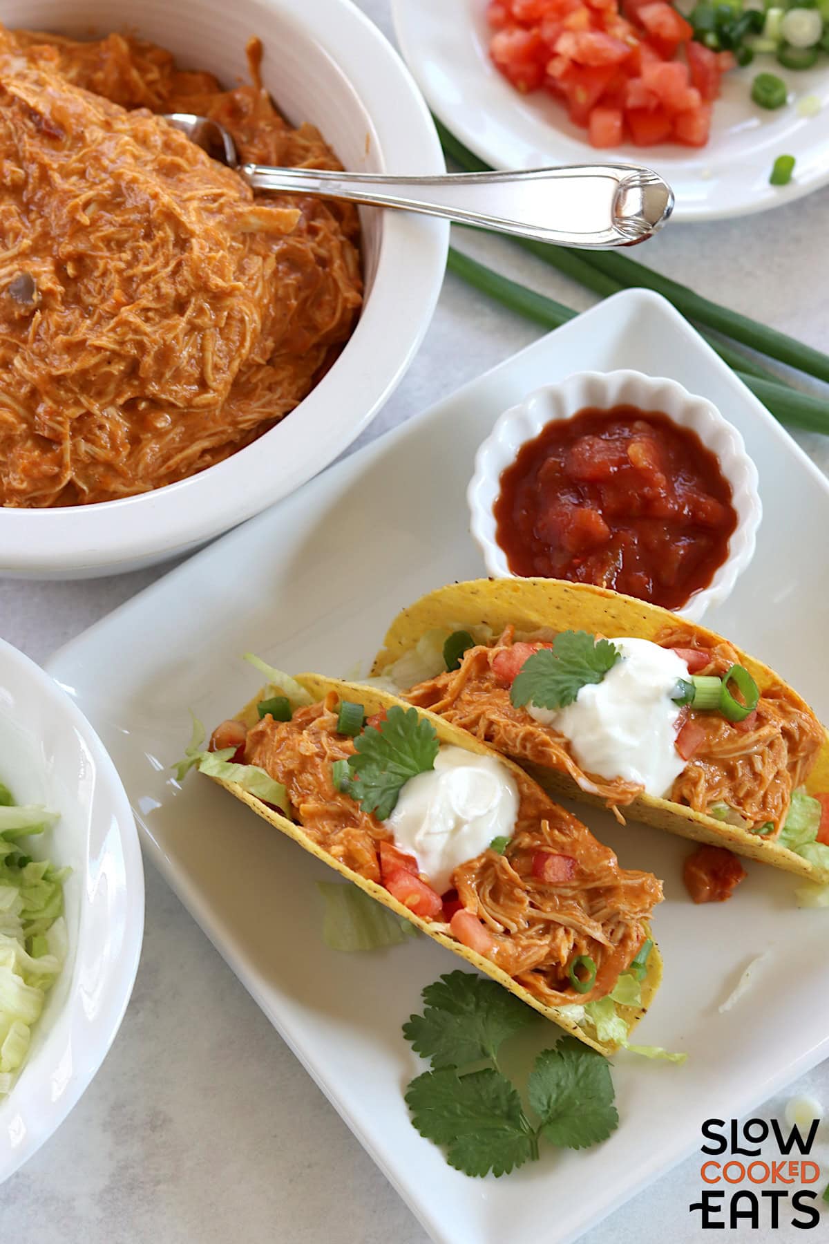 Two hard shell tacos filled with shredded queso chicken, topped with sour cream, tomatoes, and green onions on a white square plate.