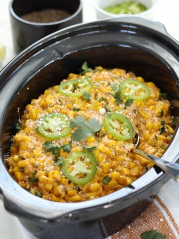 Mexican street corn crockpot recipe served in a black oval slow cooker and garnished with slices of jalapeno on top.