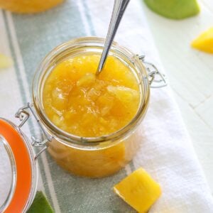 Chunky pineapple and mango crockpot jam in a jar with a spoon on a blue and white strip napkin.
