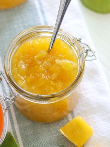 Chunky pineapple and mango crockpot jam in a jar with a spoon on a blue and white strip napkin.