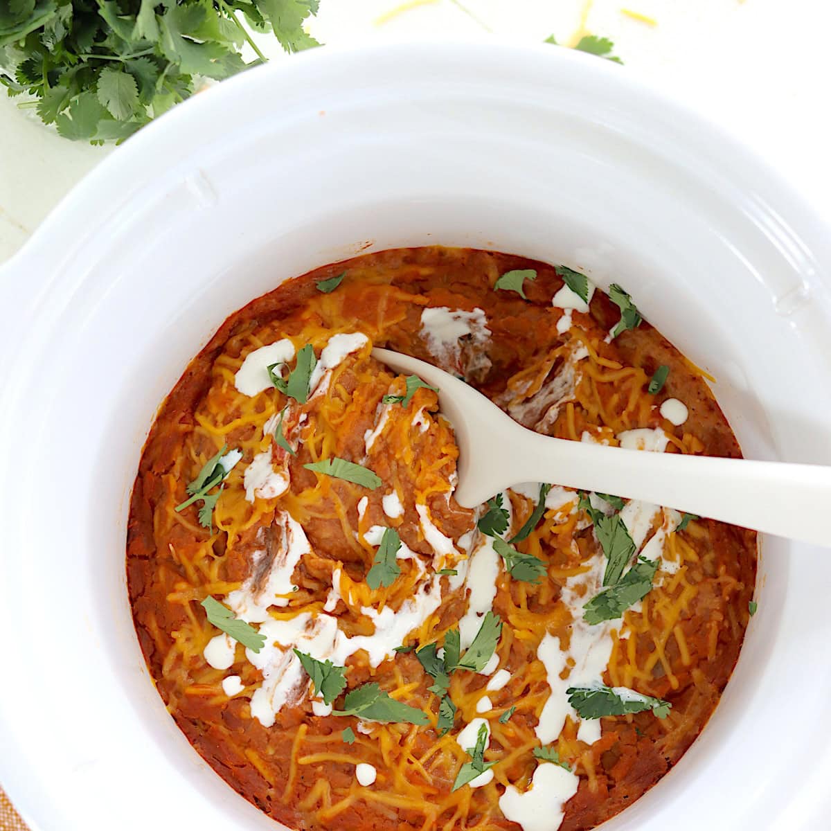 Slow cooker refried beans in a round white crockpot and a white serving spoon.