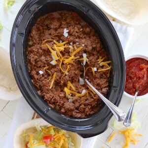 Crockpot taco meat in an oval black slow cooker and topped with diced onion and shredded cheese with a silver serving spoon.