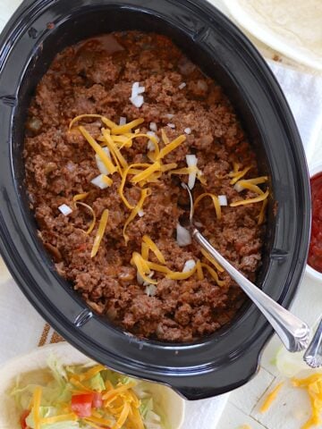Crockpot taco meat in an oval black slow cooker and topped with diced onion and shredded cheese with a silver serving spoon.
