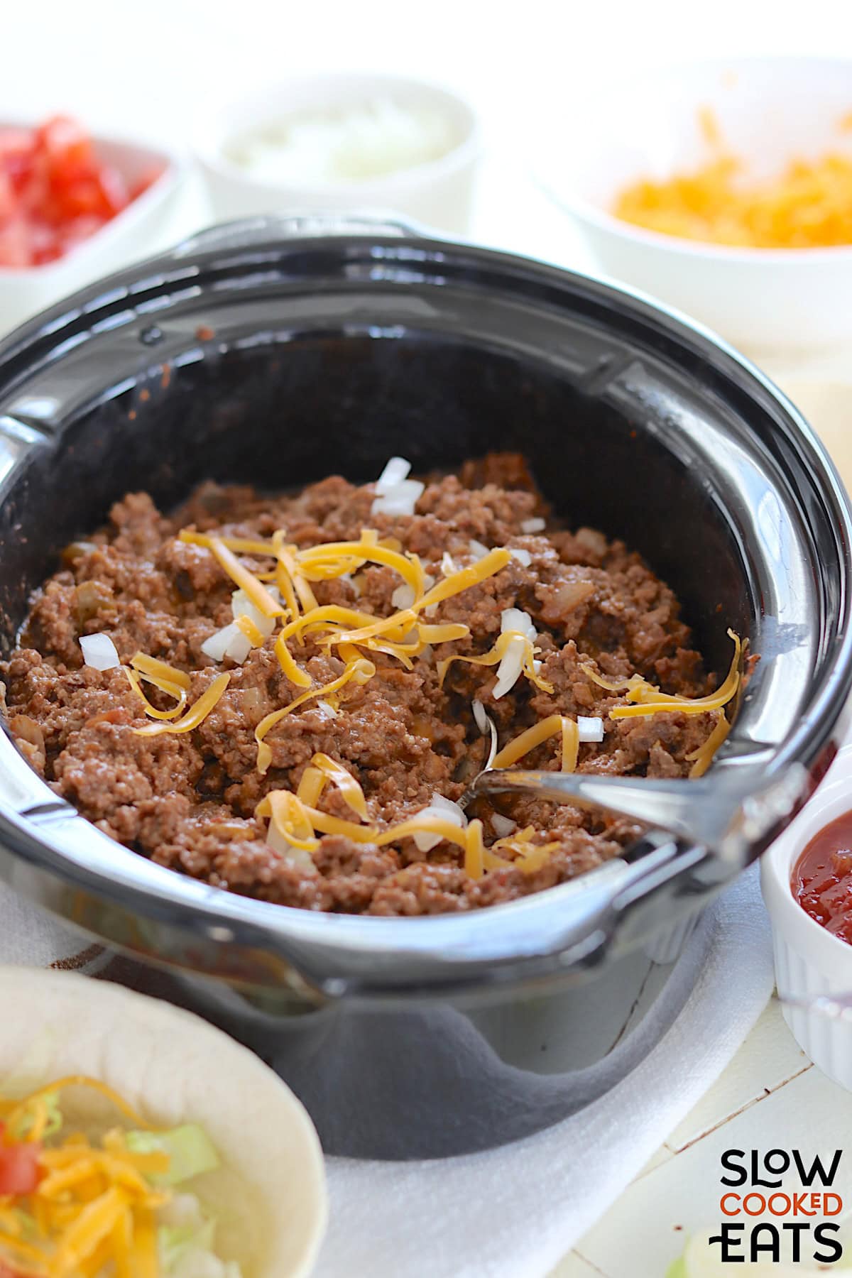 Crockpot taco meat in an oval black slow cooker, garnished with diced onion and shredded cheese with a silver serving spoon.
