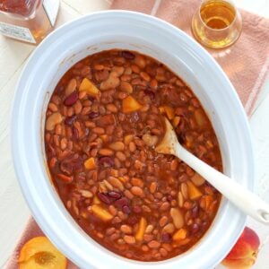 Slow cooker bourbon peach baked beans on a peach napkin with a serving spoon.