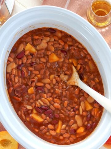 Slow cooker bourbon peach baked beans on a peach napkin with a serving spoon.