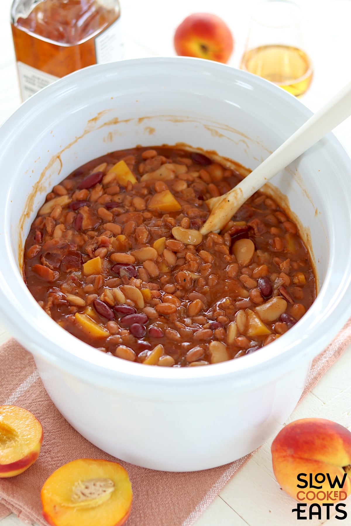 A white oval slow cooker with crockpot peach bourbon bacon baked beans with a serving spoon.