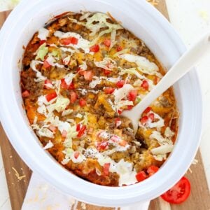Slow cooker taco pasta in a white oval crockpot insert with tomato and lettuce garnish.