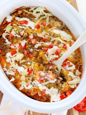 Slow cooker taco pasta in a white oval crockpot insert with tomato and lettuce garnish.