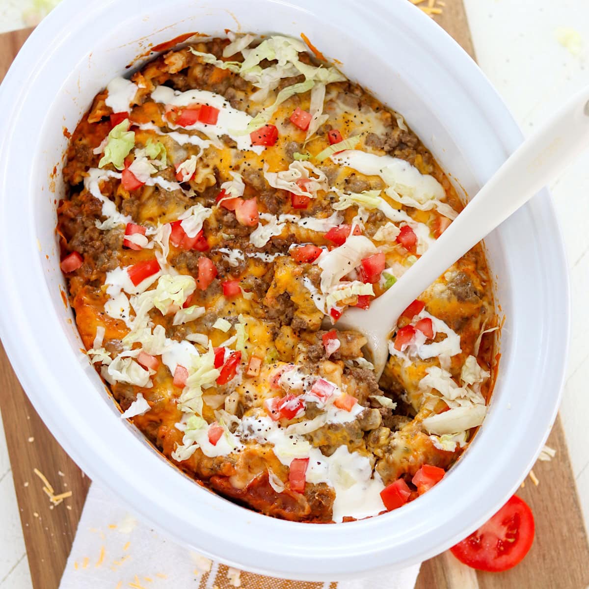 Slow cooker taco pasta in a white oval crockpot insert with tomato and lettuce garnish.